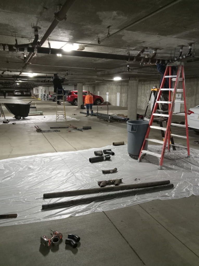 Workers repair pipes in an underground concrete parking garage, using ladders and tools spread on a plastic floor sheet.
