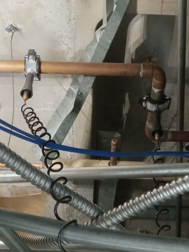 Copper piping mounted to a wall with two electrical grounding clamps attached, connected by coiled black wires.