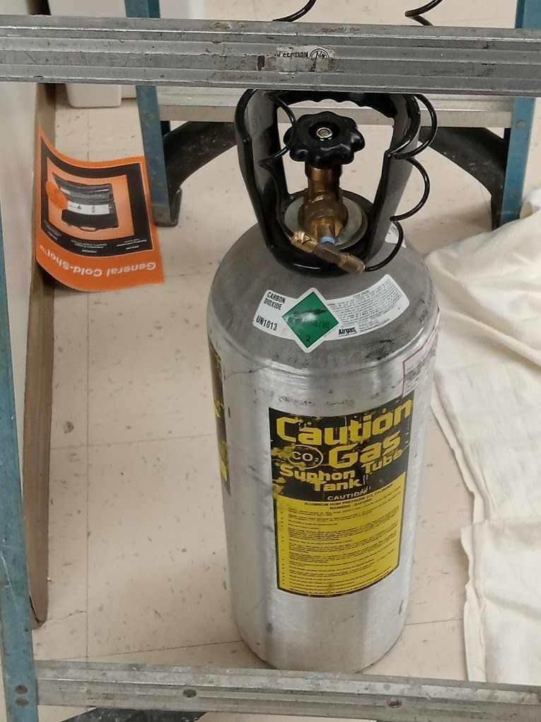 A silver compressed gas cylinder with a siphon tube warning label, standing on a floor inside a metal frame.