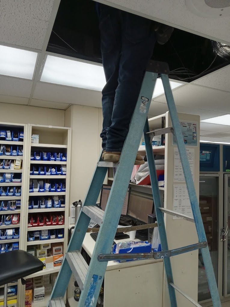 A person stands on a blue stepladder, reaching into an open ceiling panel in an office or warehouse setting.