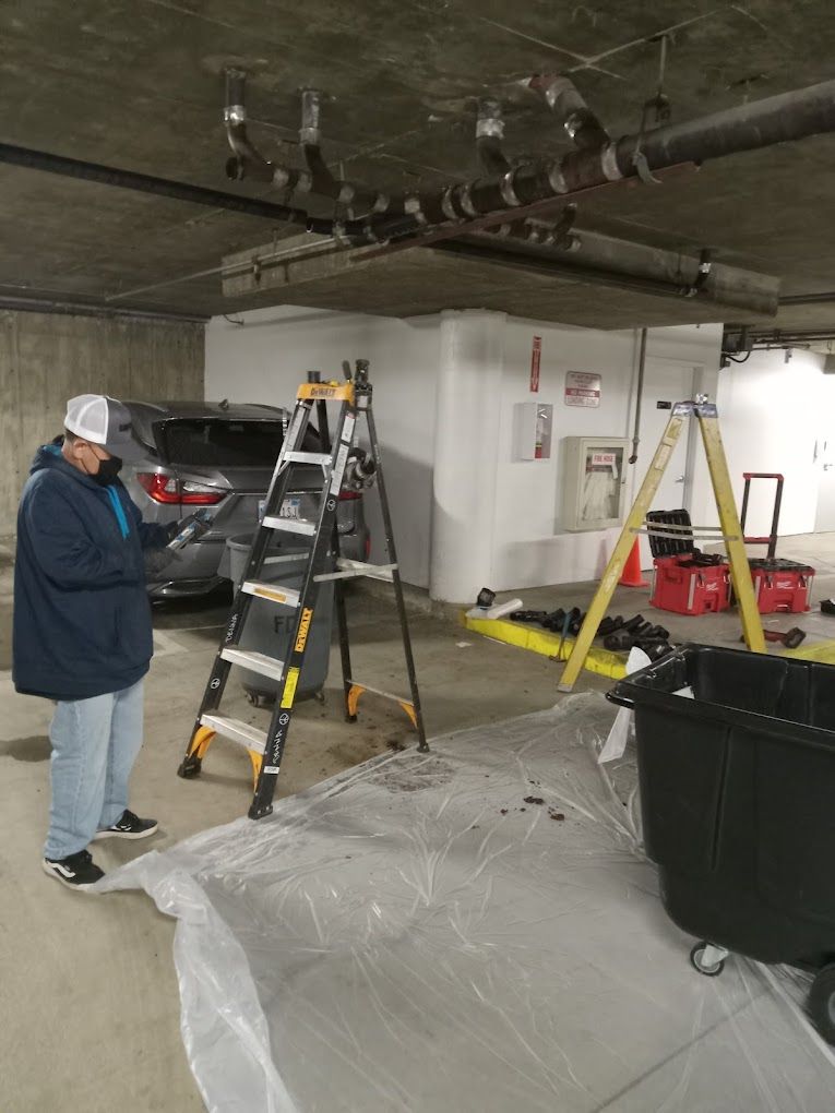 A worker stands in a parking garage near a ladder and plumbing work being done on exposed overhead pipes.