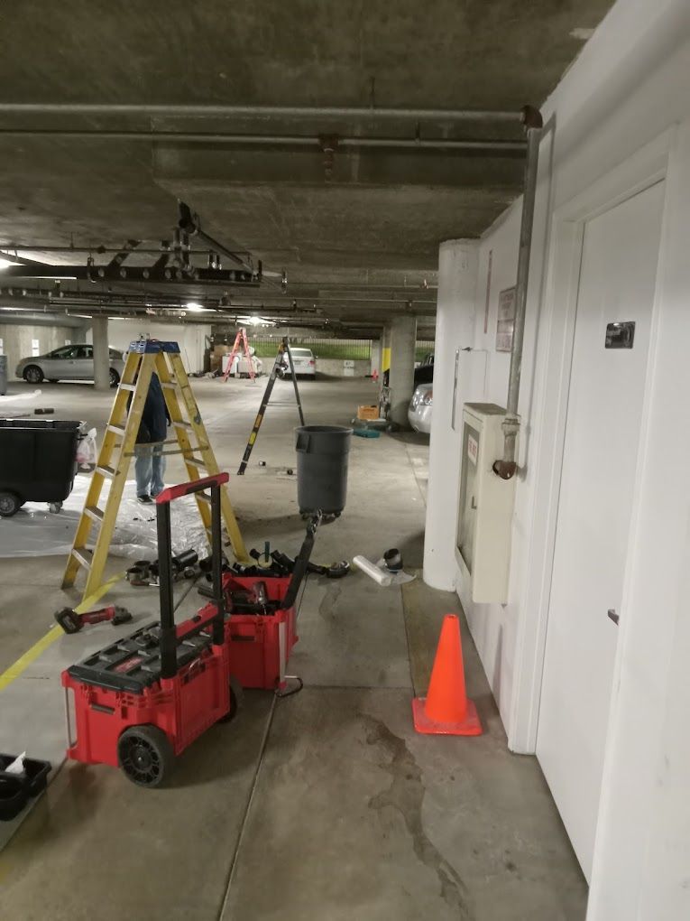 Maintenance workers using a yellow ladder and red tool chests in a parking garage with an orange safety cone.