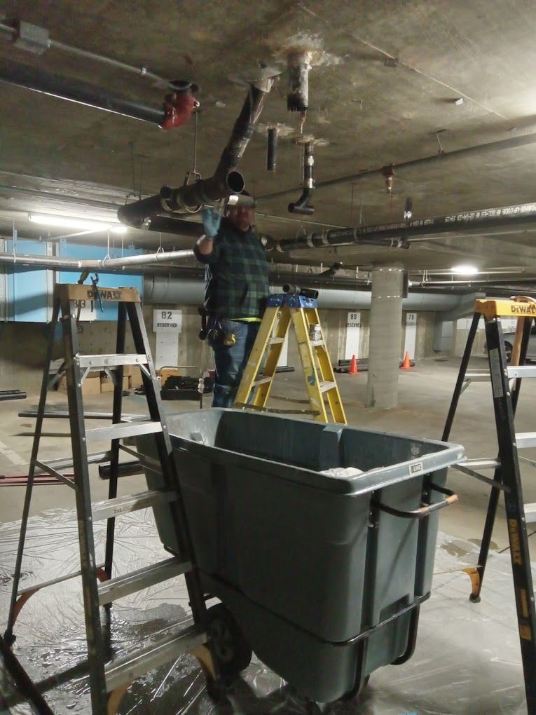 A worker on a yellow ladder repairs overhead plumbing in an industrial parking garage near a large gray utility cart.