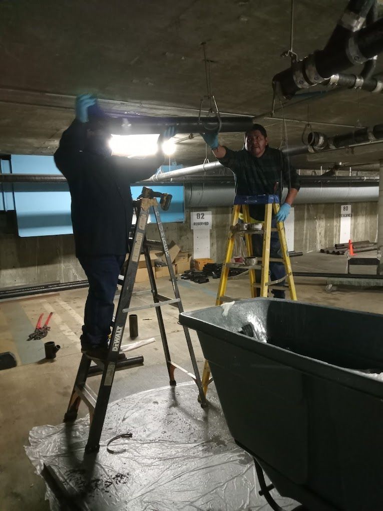 Two people on ladders repair pipes on the ceiling of a concrete parking garage near a large dark utility bin.