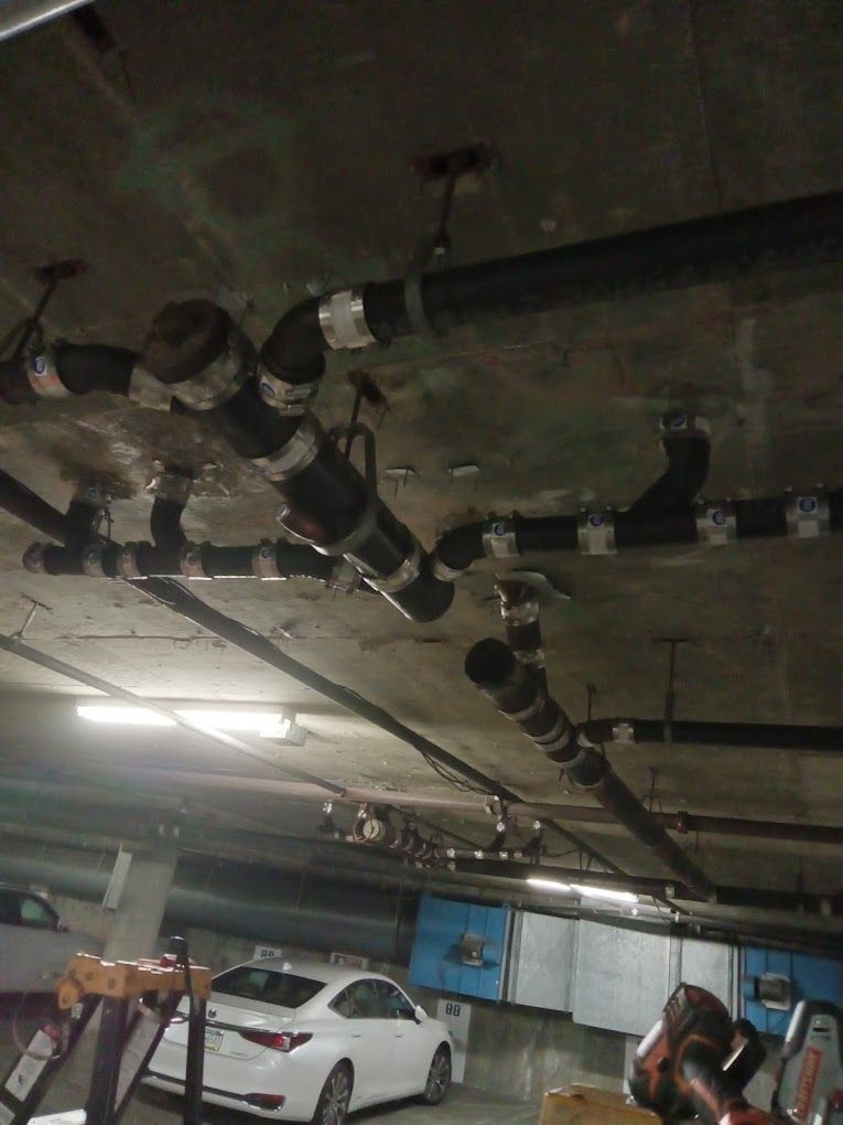 Ceiling-mounted black plumbing pipes and conduits in a parking garage with a white car in the background.