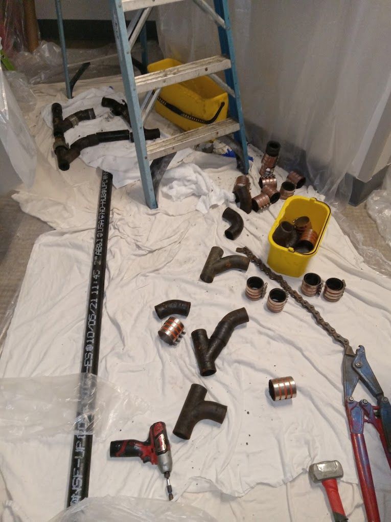 Plumbing tools and assorted black plastic and copper pipe fittings are scattered on a white drop cloth near a step ladder.