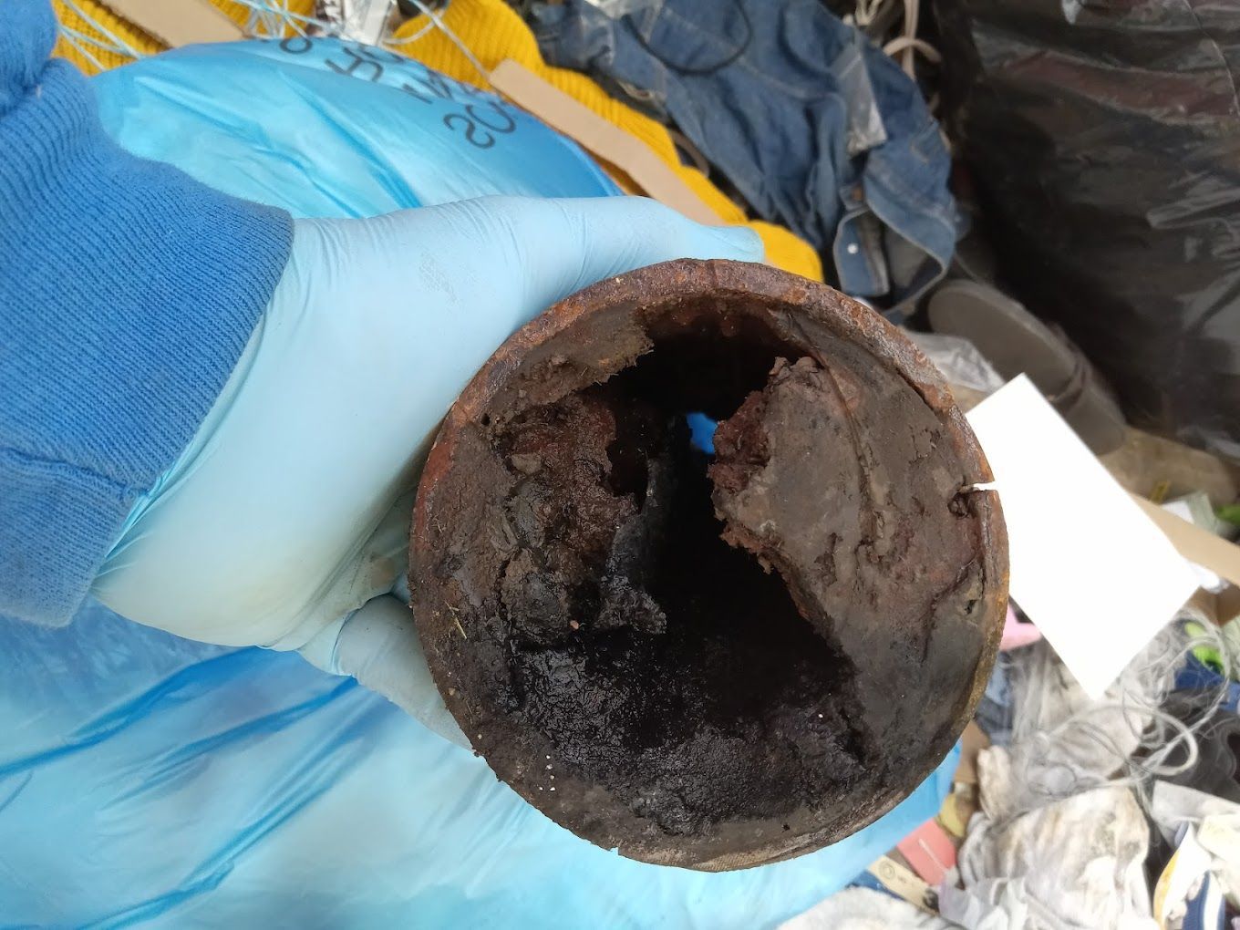 A person wearing a blue nitrile glove holds a heavily corroded metal pipe section clogged with thick, dark sludge.