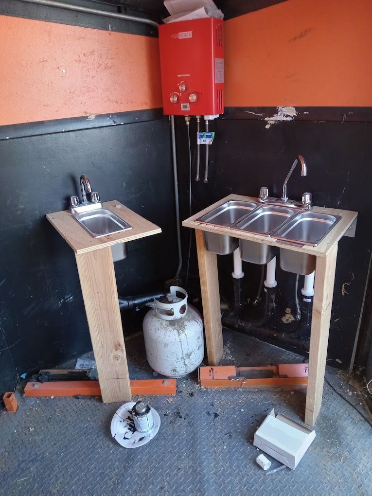 A portable sink setup with a red water heater, propane tank, and two wood-framed sinks in an orange and black walled room.