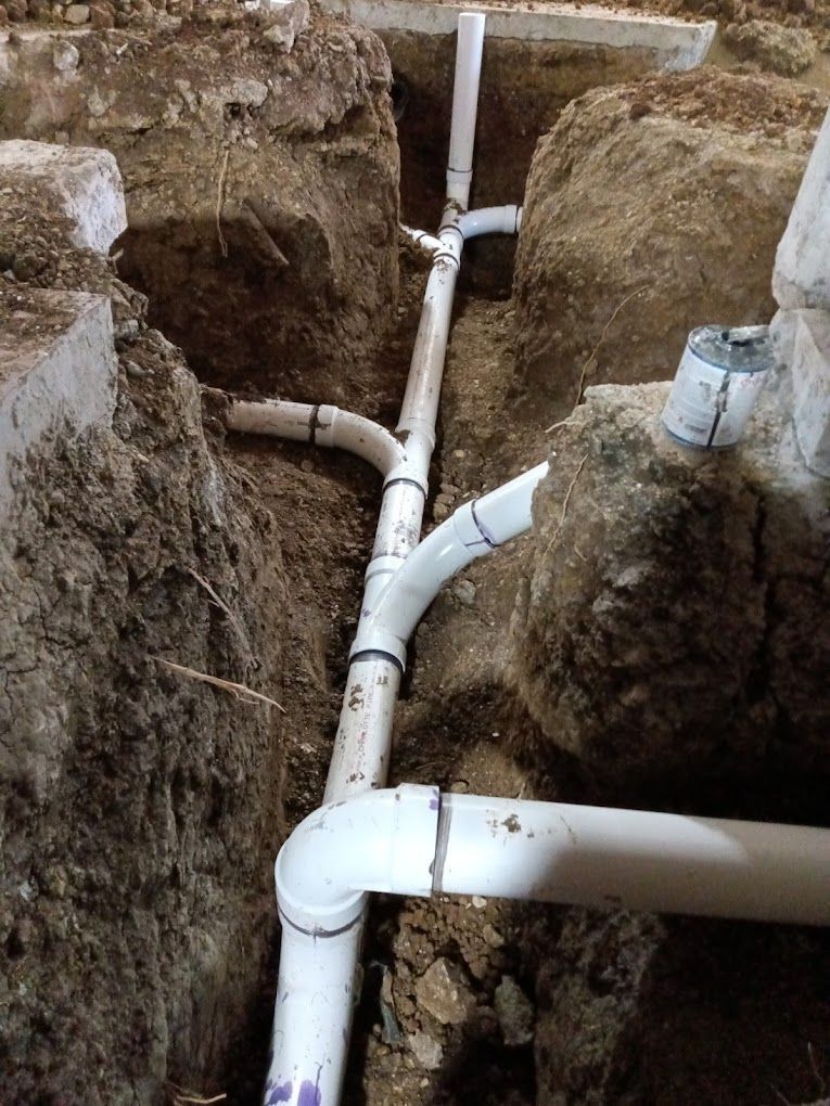 White PVC plumbing pipes installed in an open dirt trench during residential construction.