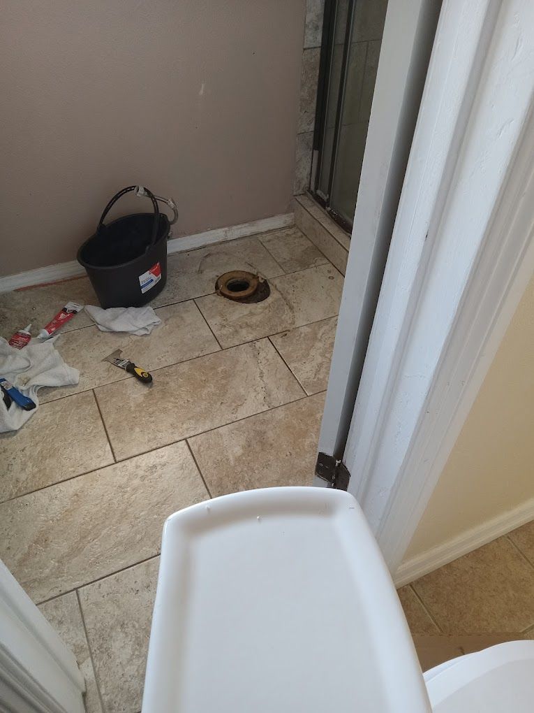 A toilet flange in a tiled bathroom floor during a renovation, with a bucket and tools nearby.