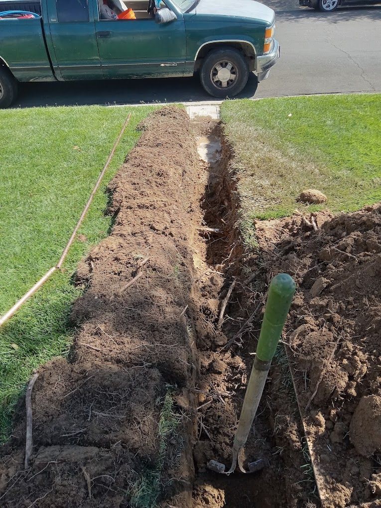 A green truck is parked on the street next to a long, narrow trench dug into a residential lawn with a shovel inside.