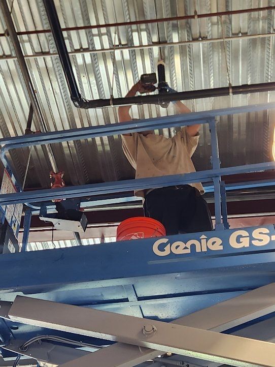 A worker in a tan shirt stands on a blue Genie scissor lift, reaching up to work on pipes installed on a metal ceiling.