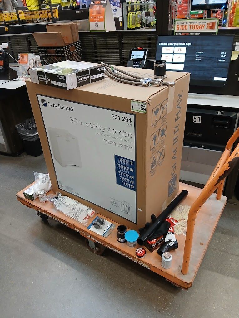 A large cardboard box containing a toilet on a flatbed shopping cart in a store aisle, surrounded by various supplies.