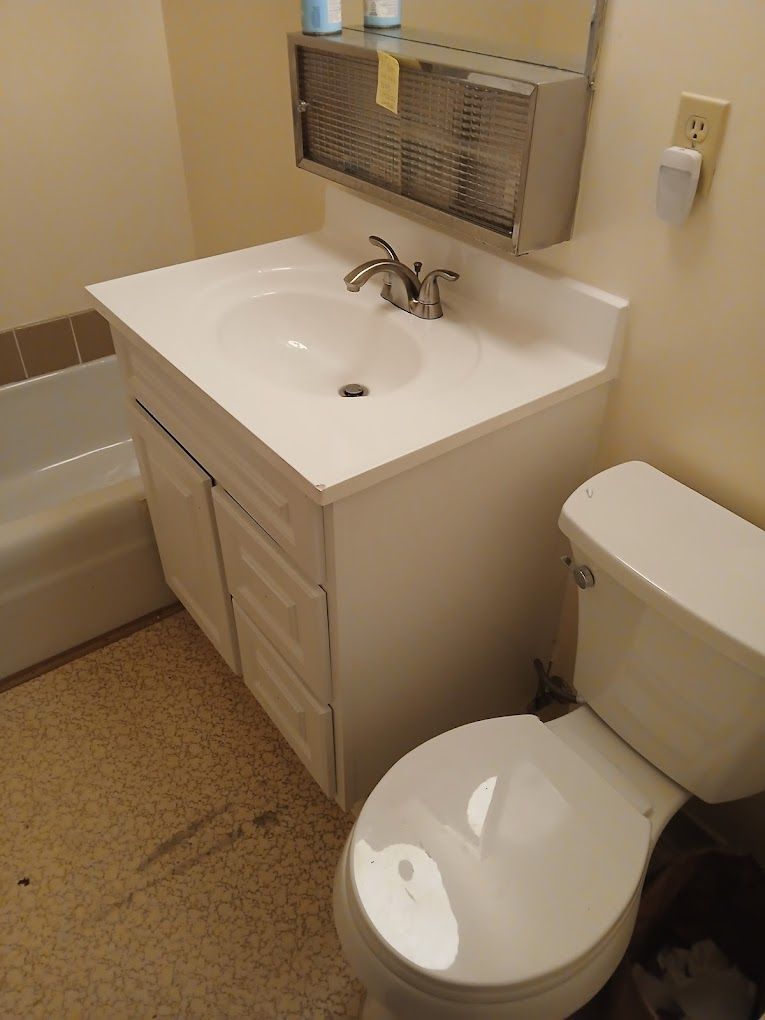 A white bathroom vanity with a sink and faucet next to a toilet and a tiled bathtub in a beige room.