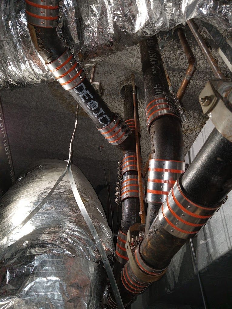 Cast iron pipes joined with rubber couplings and silver clamps running through a ceiling space next to insulated ducts.
