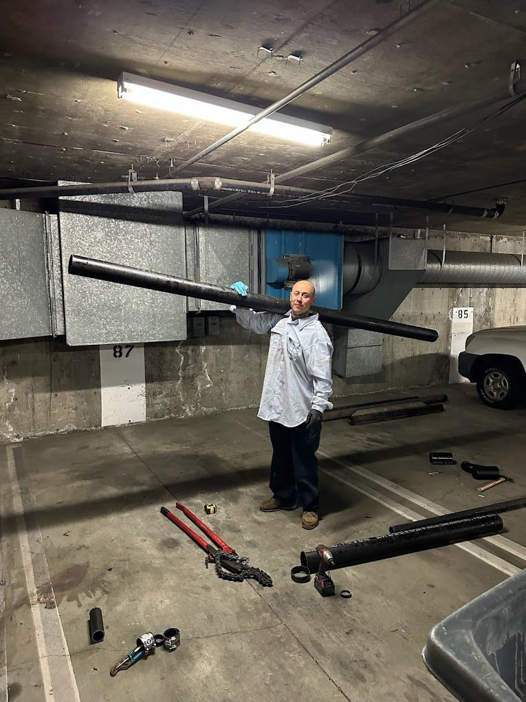 A person holds a long black pipe horizontally across their shoulders in an indoor parking garage with tools on the floor.