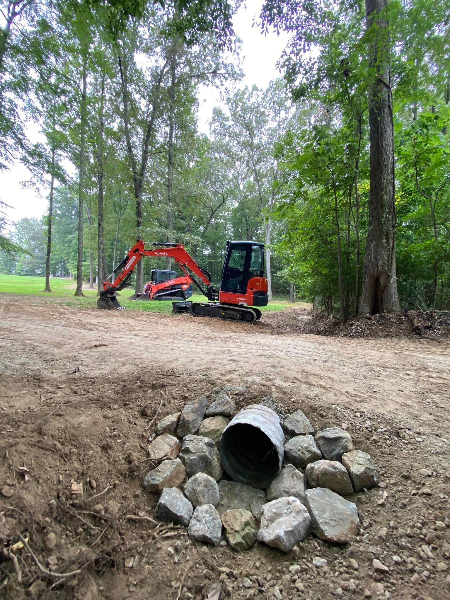 A red excavator works on a gravel path near a drainage pipe framed by rocks in a wooded area.
