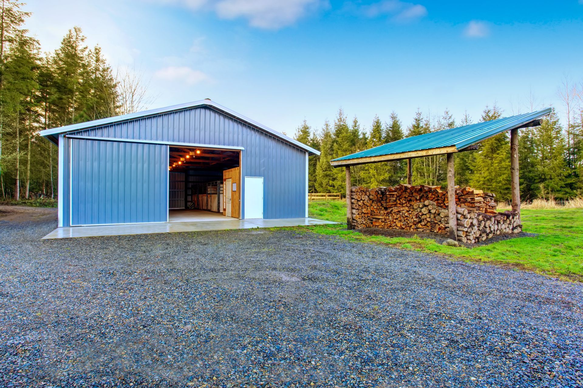 A blue metal barn with a sliding door open and an adjacent firewood shed on a gravel lot near a forest.