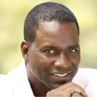 Close-up of a smiling Black man with short hair, resting his chin on his hand. Outdoors, soft lighting.