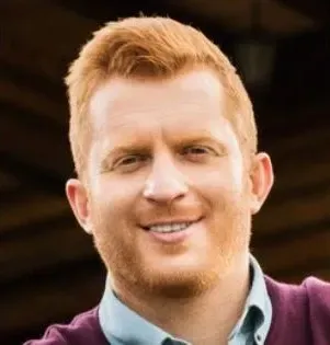 Man with red hair and beard smiling; wearing a blue shirt and burgundy sweater.
