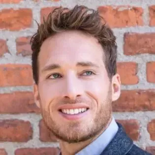 Man with fair skin, light eyes, and a beard smiles against a red brick wall.
