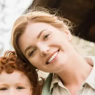 Woman with blonde hair smiles, head next to a child with red hair. Outdoors, natural light.