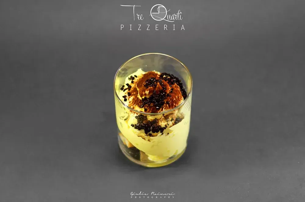 Dessert in un bicchiere con crema gialla e guarnizioni scure, su una superficie grigia.