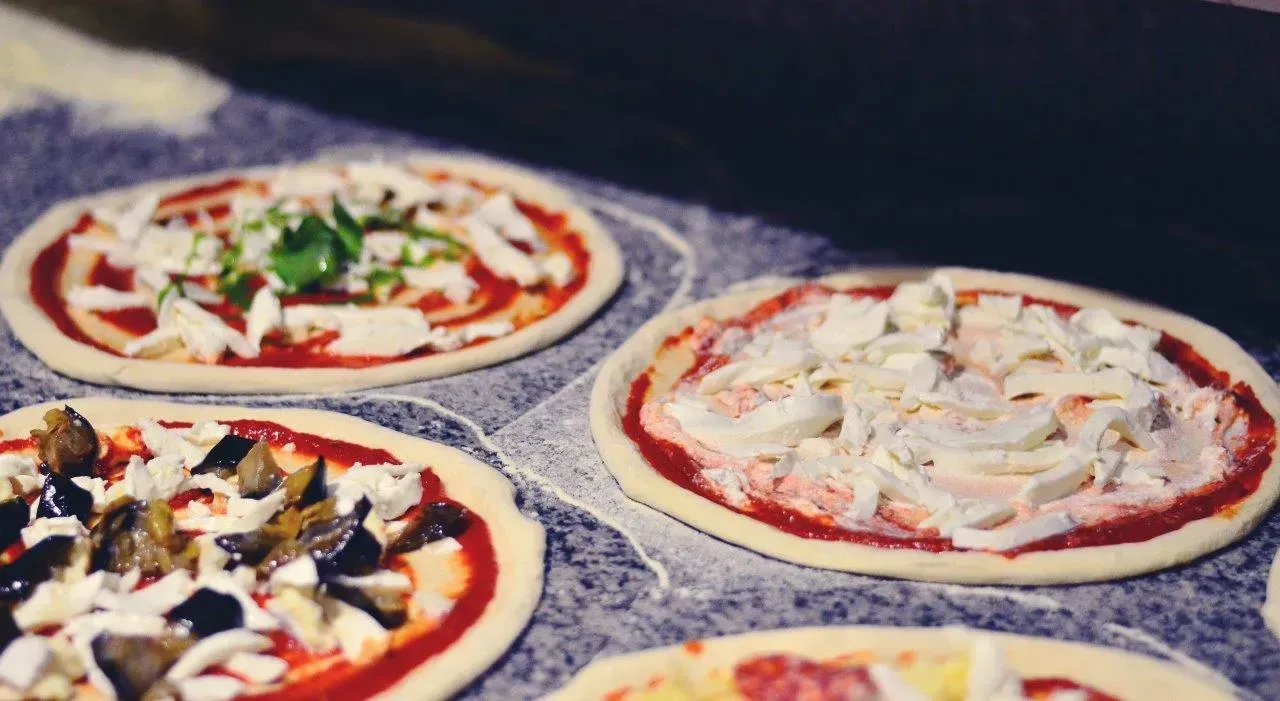 Quattro pizze crude con condimenti diversi, disposte su una superficie grigia.