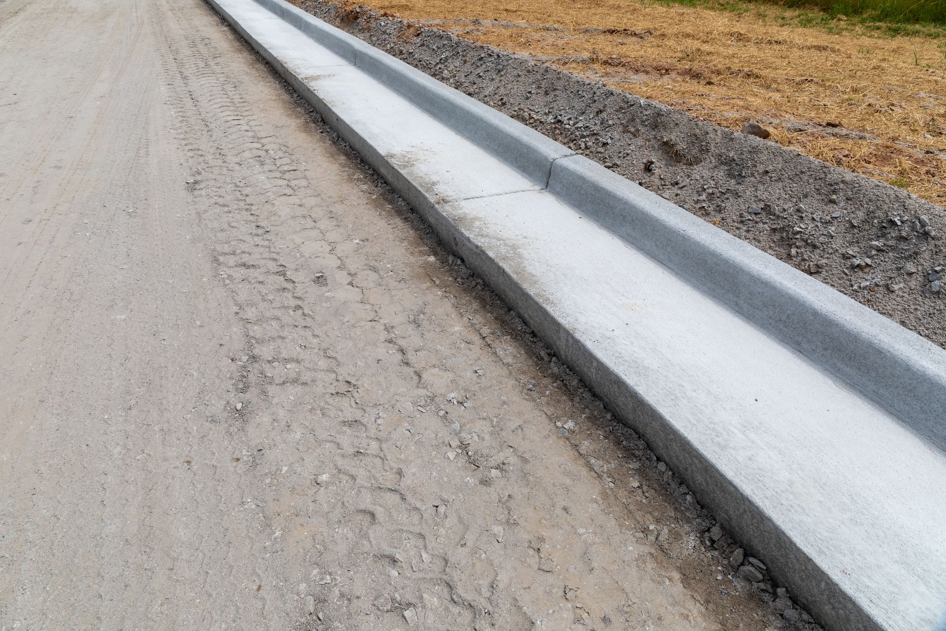 Concrete curb and gutter along a gravel road, with dirt and grass in the background.