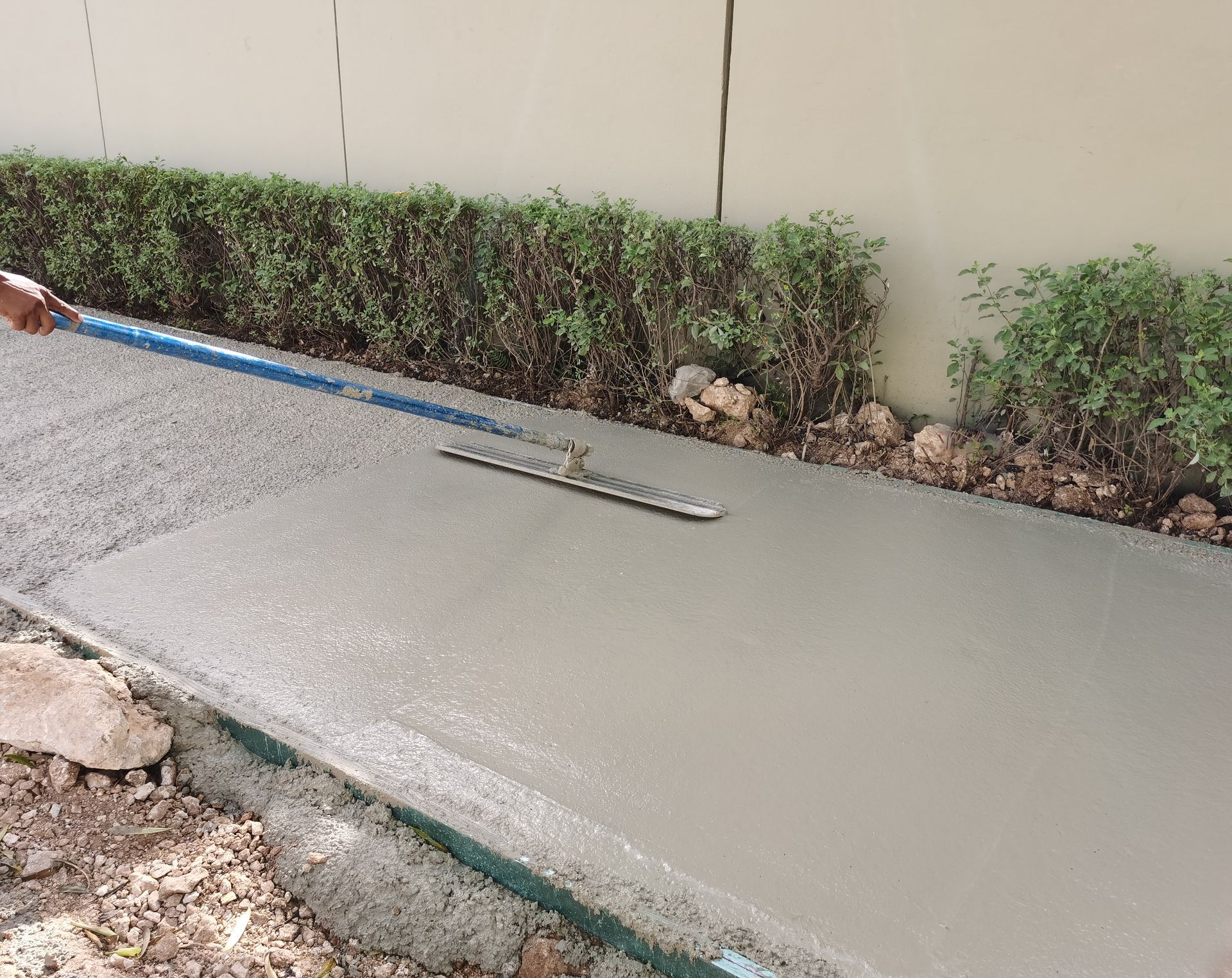 Person using a screed to level wet concrete in a walkway, next to a hedge and a wall.