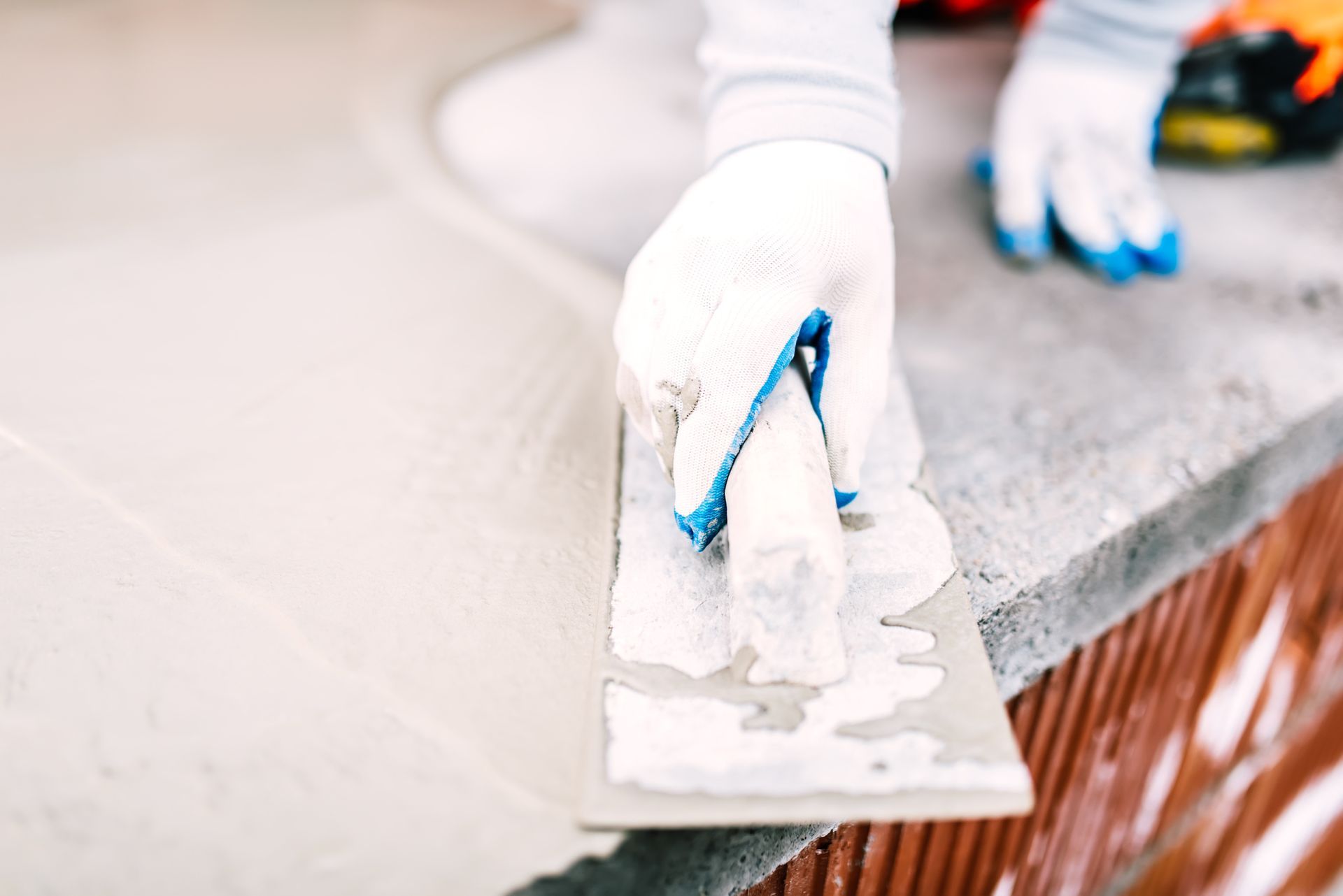 A gloved hand uses a trowel to smooth wet cement on a brick surface.