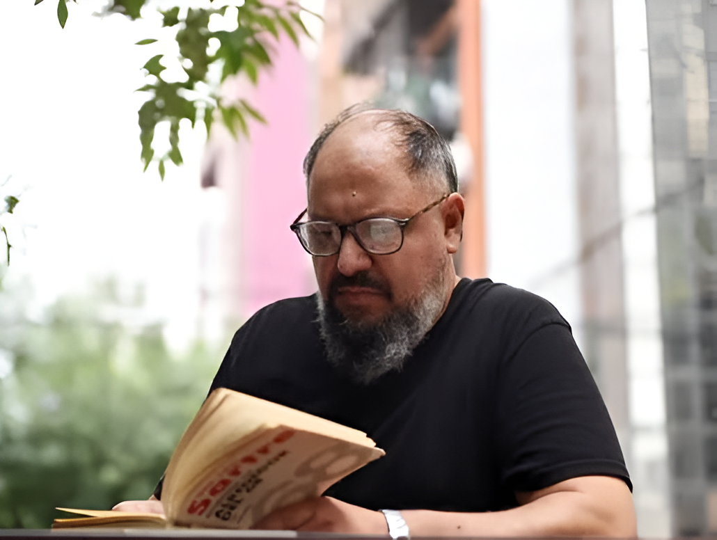 Hombre con gafas y barba leyendo un libro al aire libre; fondo borroso de edificios y follaje.