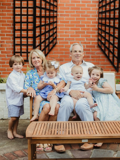 Family seated outdoors, smiling. Includes older adults holding young children in front of a brick wall and wooden lattice.