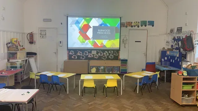 A classroom with tables and chairs and a projector screen on the wall.