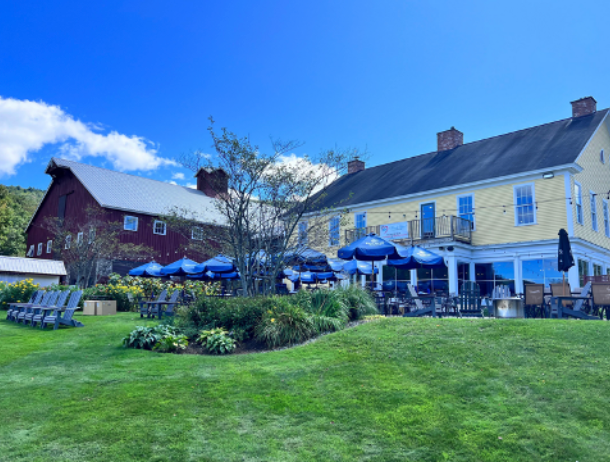 Yellow building with blue umbrellas, red barn, and green lawn under a blue sky.