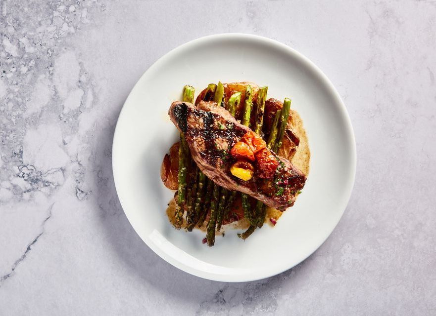 Grilled steak and asparagus over mashed potatoes on a white plate, set on a marble surface.