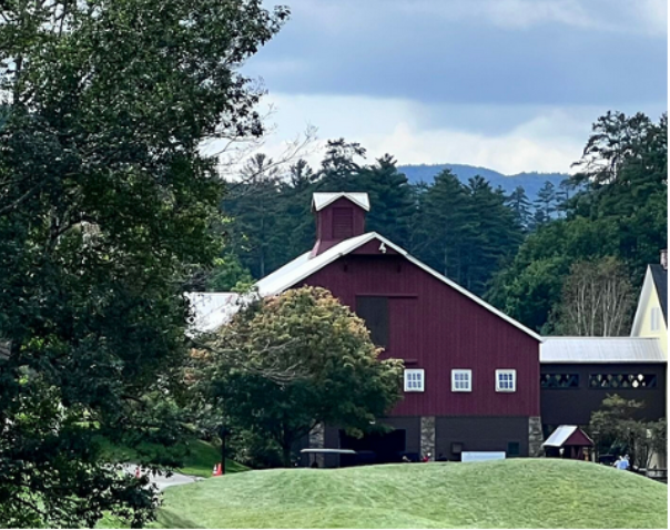 Red barn with white roof and cupola, set among trees and green grass, mountains in the background.