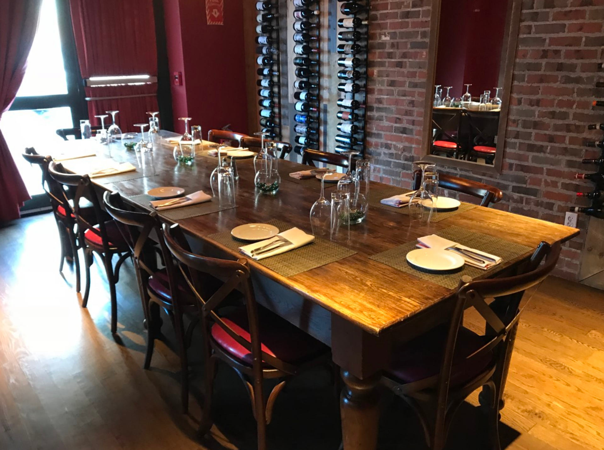 Long wooden dining table set with glassware and place settings. Red chairs. Wine rack on brick wall.