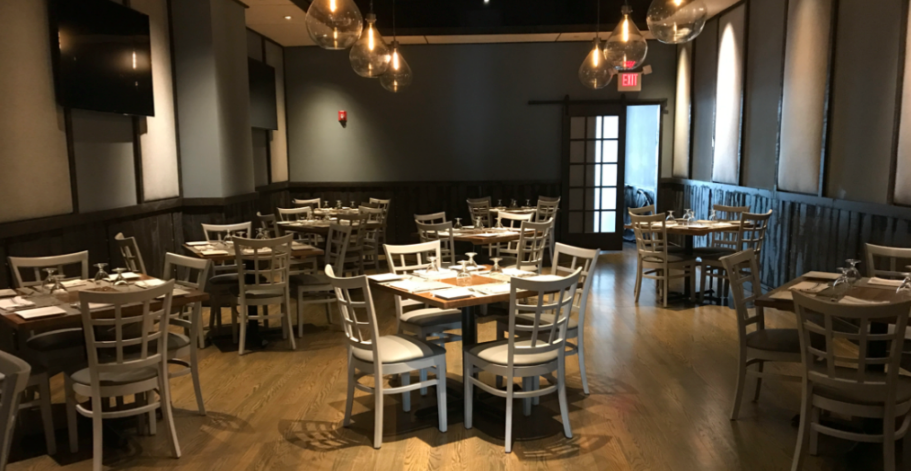 Empty restaurant dining room with tables, chairs, and hanging lights; wood floor and gray walls.