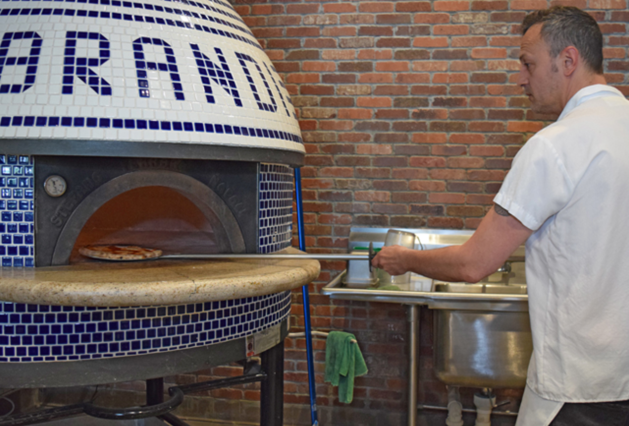 Chef taking pizza from a brick oven, inside a restaurant kitchen. Oven has blue and white tiles.