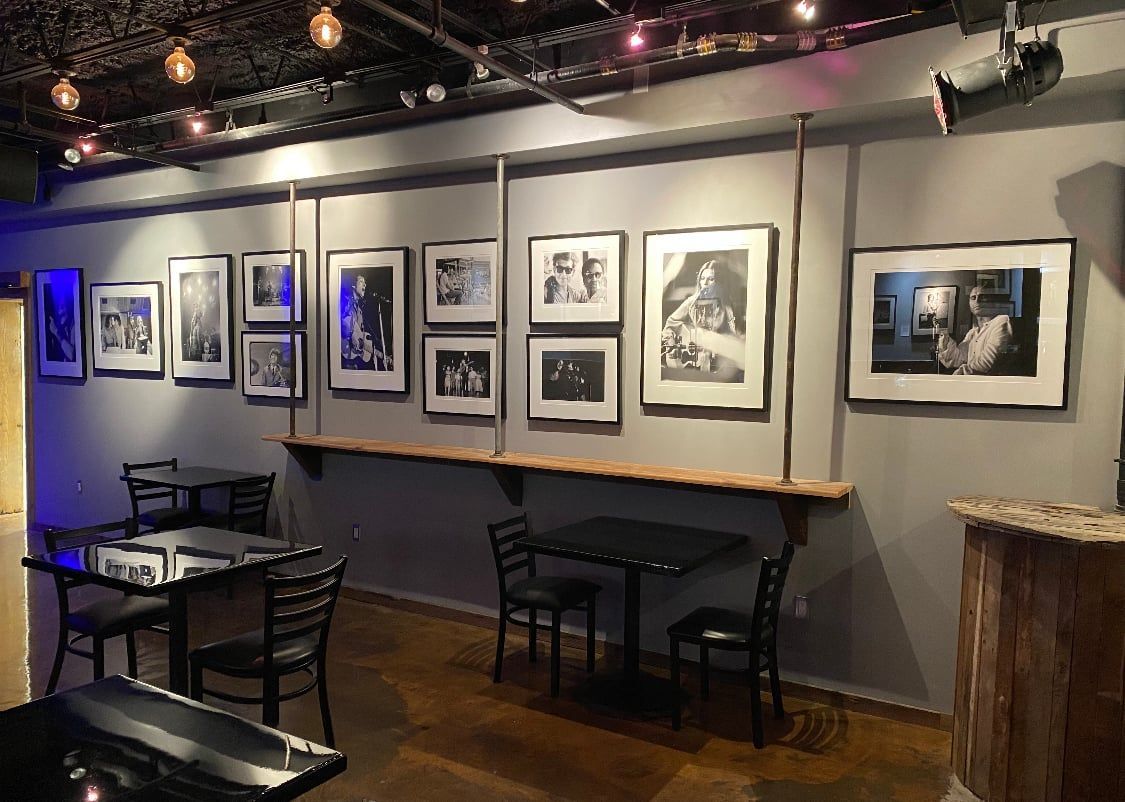 Interior of a gallery or bar. Black and white photos line the wall above a counter. Tables and chairs sit in front.