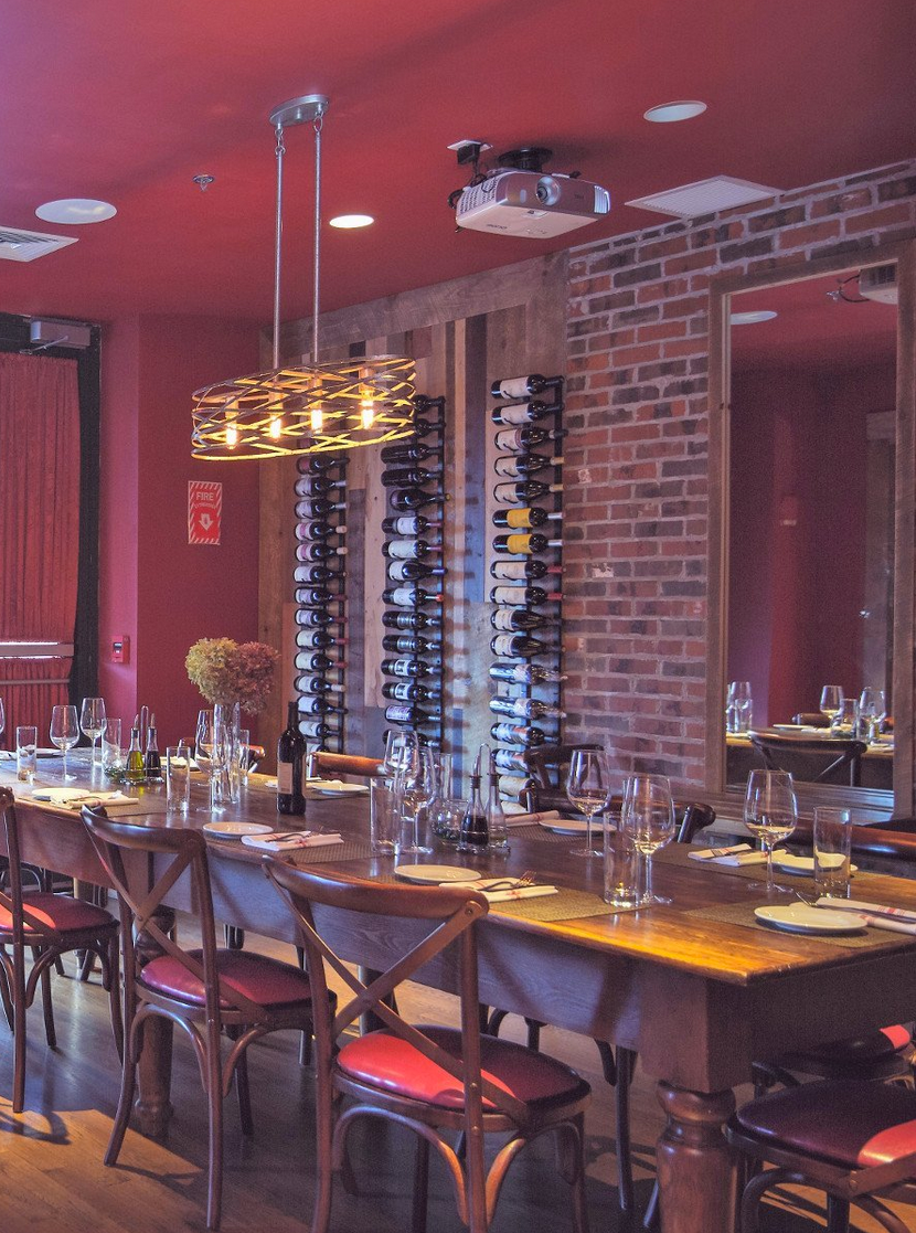 Restaurant dining room with long wooden table, red chairs, brick wall, and wine racks.