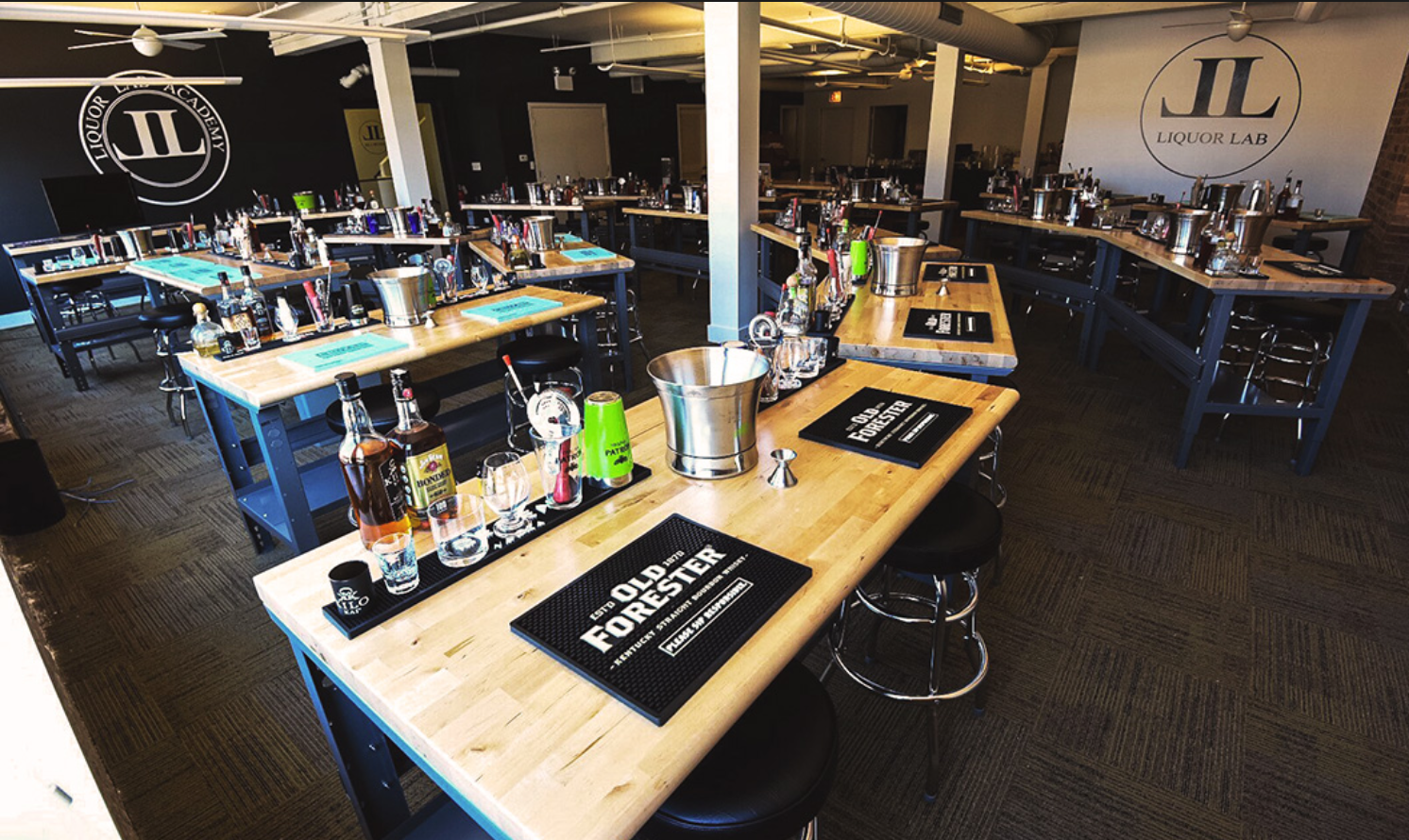 Cocktail-making class: multiple stations with bar tools, liquor bottles, and a logo on a black wall.