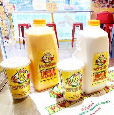 Papaya King drinks: yellow & white liquids in jugs and cups with a smiling hot dog logo on a countertop.