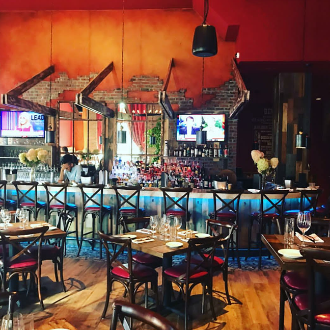 Interior of a restaurant with a bar, tables, and red seating. Brick wall, TVs, and bar lighting.