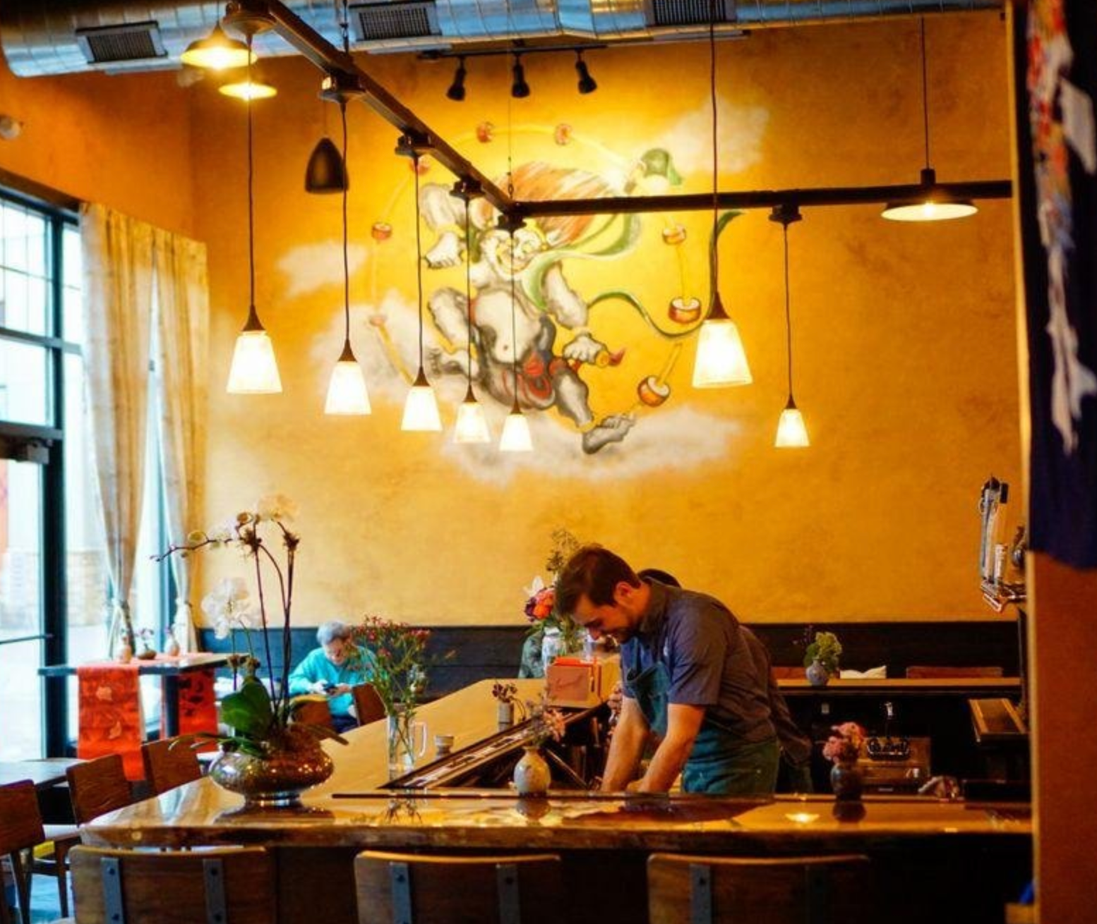Bartender working behind a counter with a mural on the wall, warm lighting, restaurant setting.