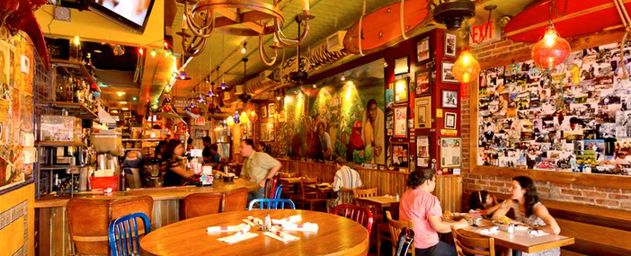 Interior of a brightly lit, casual restaurant; people eating, framed photos on walls, and a bar.