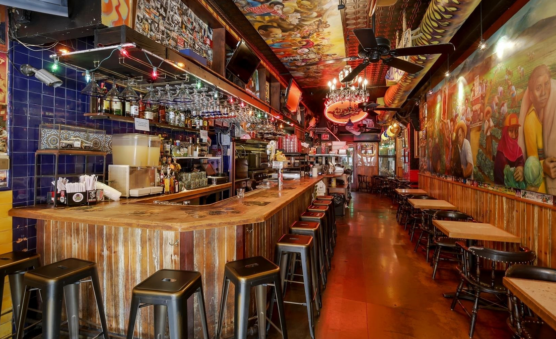 Bar interior with rustic wood bar and stools, colorful murals, and dim lighting.