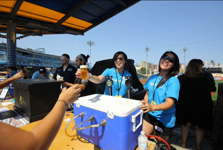 Two women serving beer at a concession stand. Sunny day, blue t-shirts, smiles. Stadium in the background.
