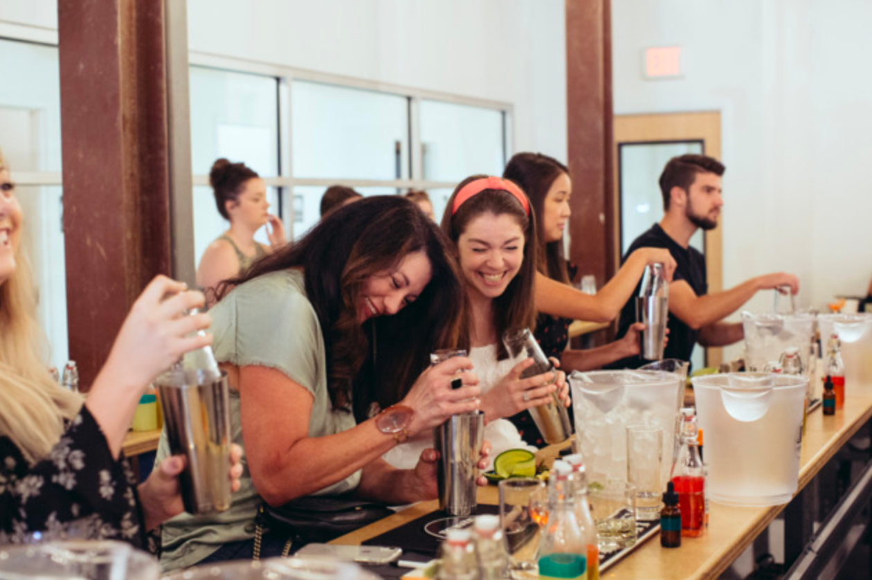 People laughing while shaking cocktails behind a bar.
